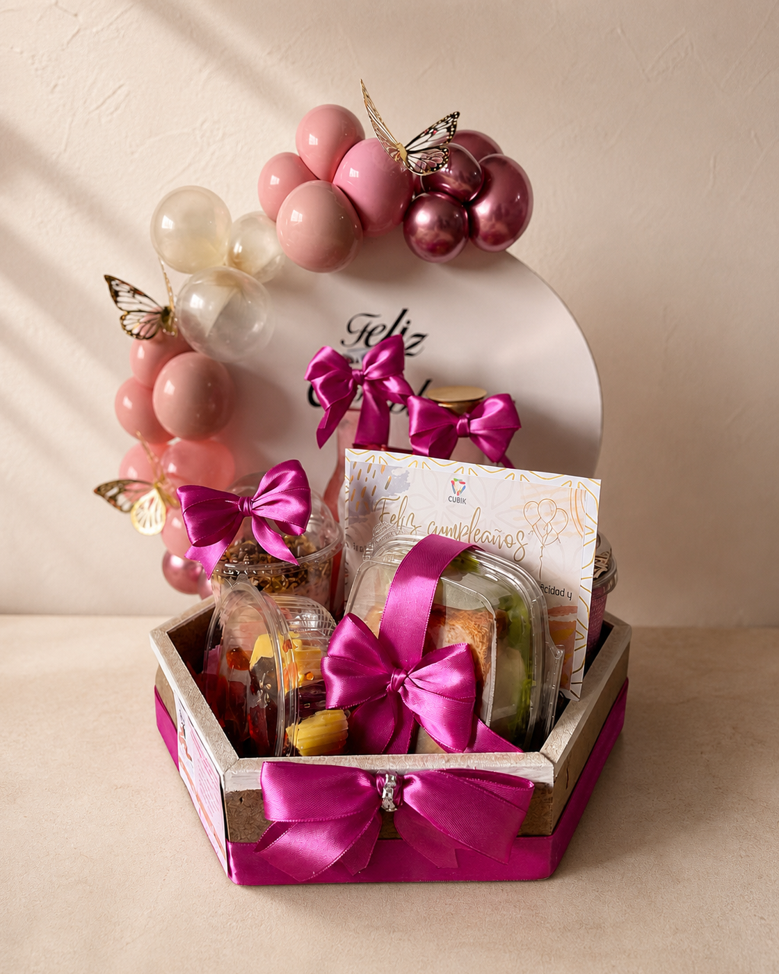 Ancheta de cumpleaños hexagonal con arco de globos rosa y granate, mariposas doradas y bandejas con fruta y aperitivos