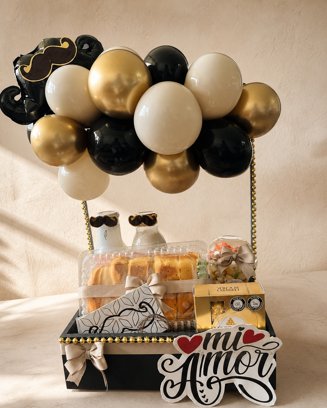 Bandeja de regalo en negro y dorado con globos metalizados, dulces y cartel de mi amor