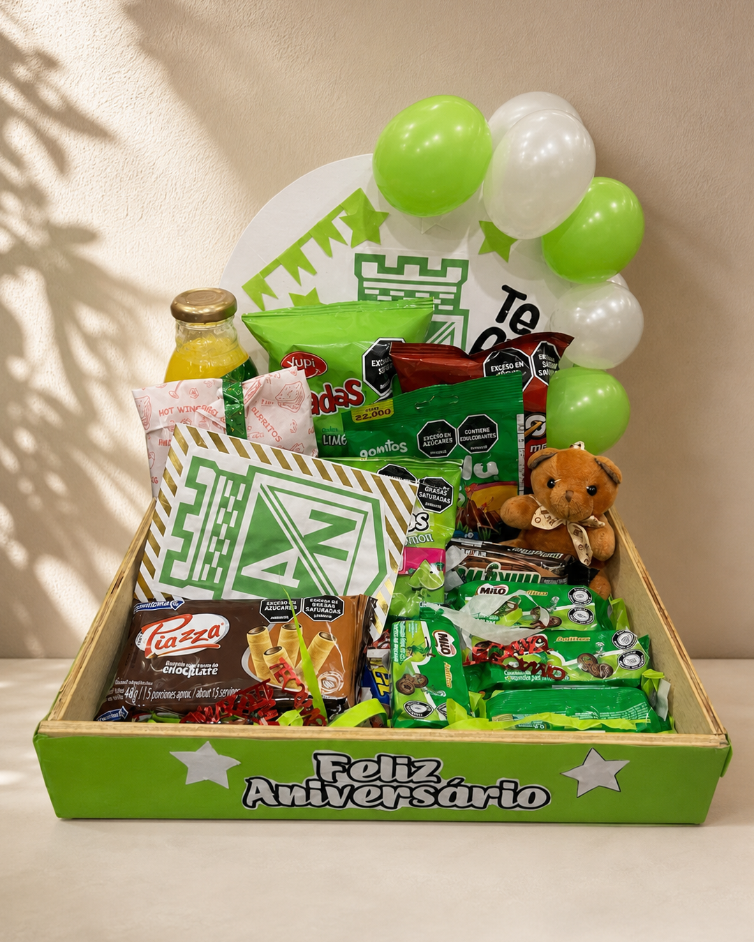 Ancheta de aniversario en caja verde con globos verdes y blancos, snacks, chocolates y peluche de oso