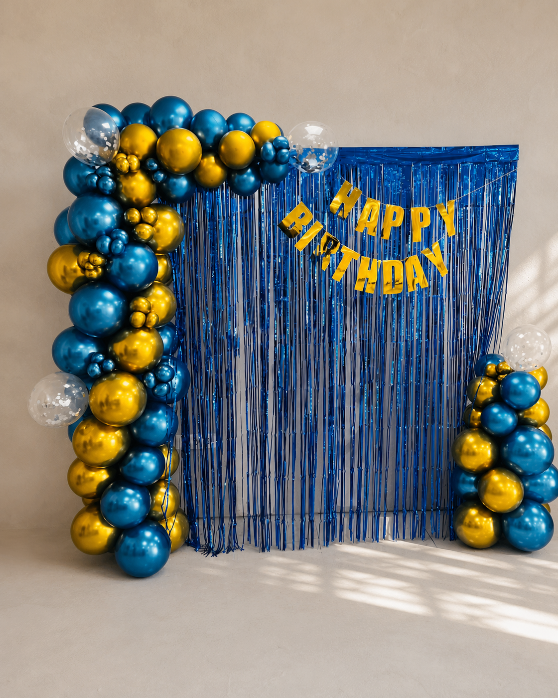 Photocall de cumpleaños con arco de globos en azul y dorado sobre cortina de flecos